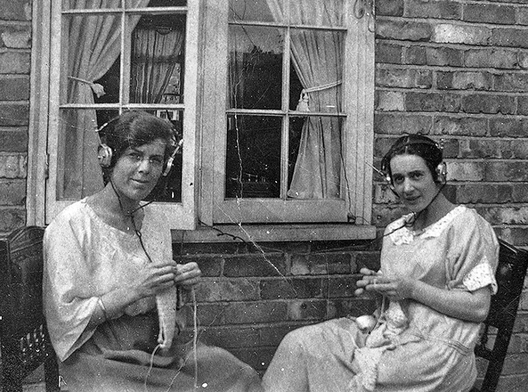 Two women sitting outside knitting and listening to the radio through headphones, c1920s