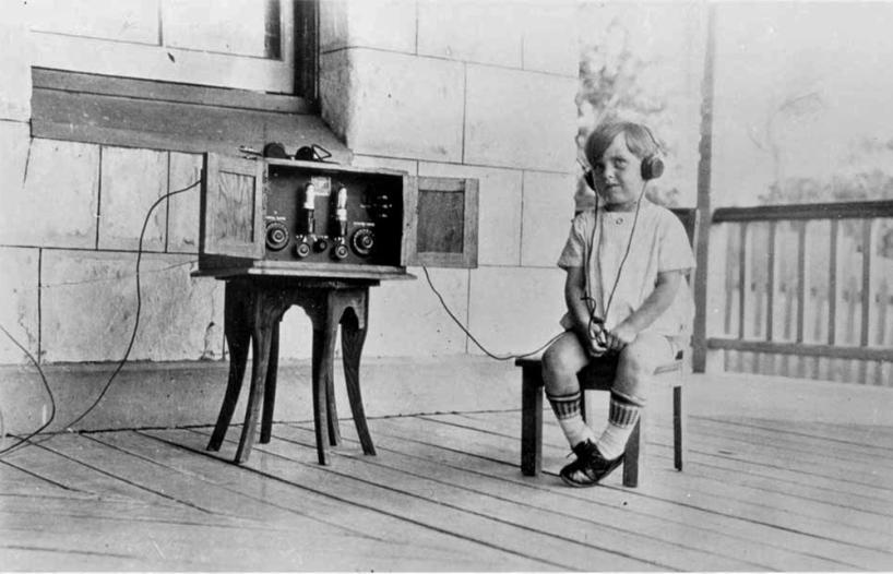 A child sitting on a stool and wearing headphones listening to the radio, c1930s.