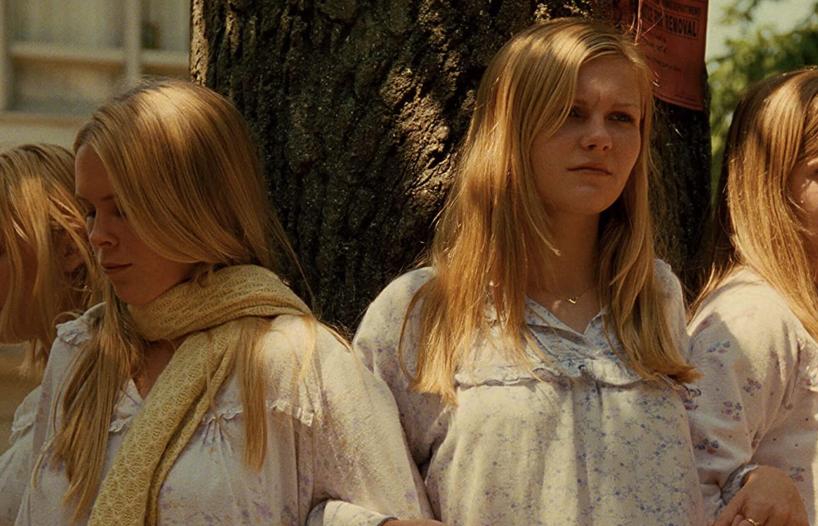 Four young women with long blonde hair wearing pale nighties and standing outside around a tree during the daytime, with mournful expressions