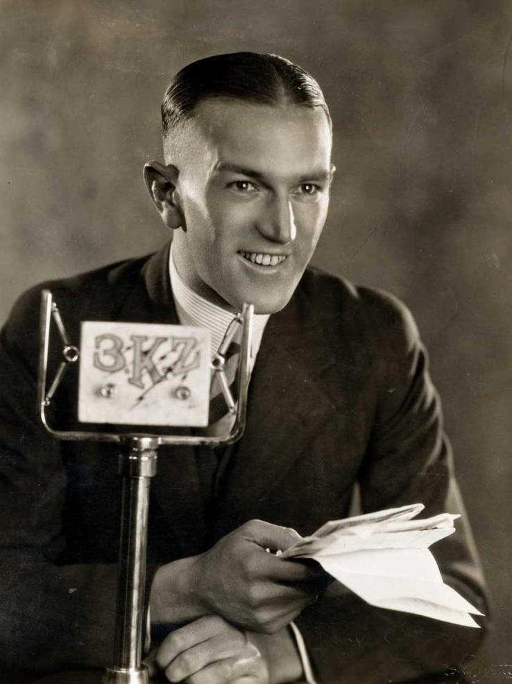 A man holding some papers and smiling. He is seated behind a 3KZ radio microphone. 