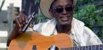A middle aged African American man wearing a hat and sunglasses man holding a small bottle in one hand and a guitar. 