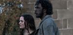 Aisling Franciosi and Baykali Ganambarr are standing outside in front of a wall in a scene from the film The Nightingale. They are looking pensively off camera