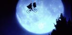 A large moon in the night sky behind a silhouette of a child riding a flying bicycle with a basket on the handle bars. 