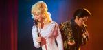 A woman dressed like Dolly Parton in a white dress and blonde wig sings into a microphone next to someone dressed as Elvis Presley
