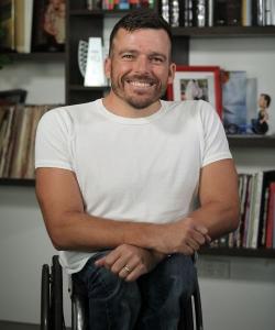 A portrait of wheelchair racer Kurt Fearnley smiling for the camera. He has bookshelves filled with books behind him.
