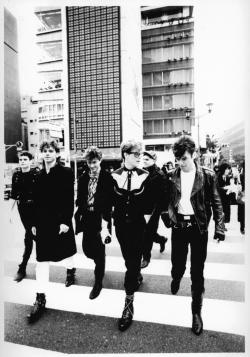 Publicity image of the six band members of INXS walking across a pedestrian crossing with tall buildings in the background.