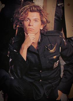 Singer Michael Hutchence kneeling on the ground and resting his chin in his hand and looking directly at camera, wearing a black military style jacket.