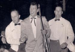 Col Joye holding his guitar being led away by two men in white jackets and black bow ties.
