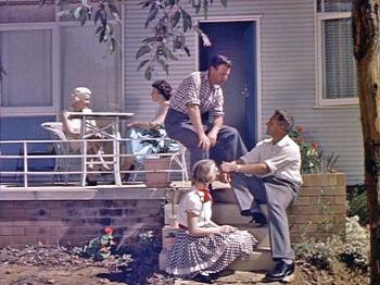 A group of adults and a teenage girl sitting out the front of a suburban Australian house in the late 1950s.