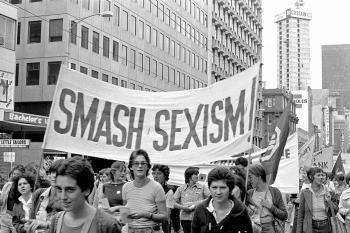 A women's liberation march on the streets of Sydney. Hundreds of women on the street. Some are holding a large sign that says 'Smash sexism'.
