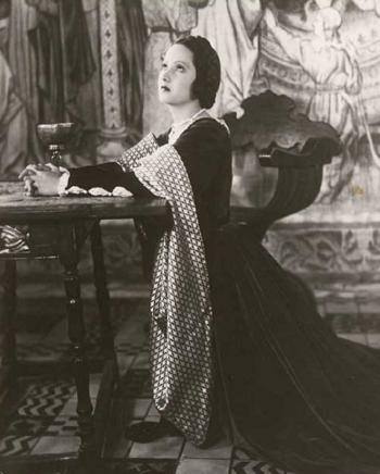 Merle Oberon as Anne Boleyn kneeling at a table with her hands clasped and her eyes uplifted