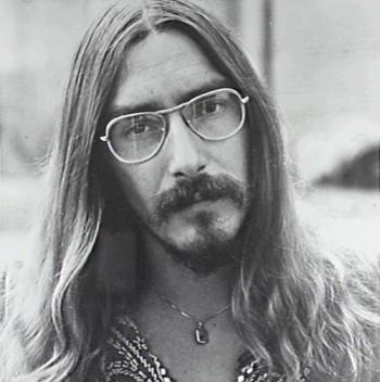 Head and shoulders close up of singer Jeff St John wearing glasses, a necklace with pendant and embroidered shirt, looking directly at camera with his head slightly tilted to his right. The image is dated around the late 1970s.