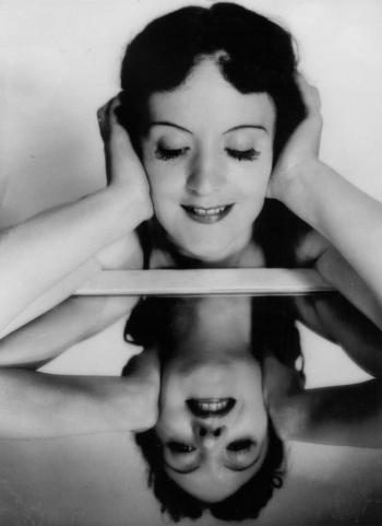 A smiling Diana Du Cane holds her head and gazes downwards at her reflection in a mirror