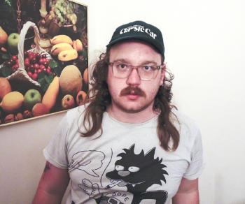 Tom Martin wearing baseball cap and T shirt with a black cat smoking. Poster of fruit and vegetables on the wall behind him.