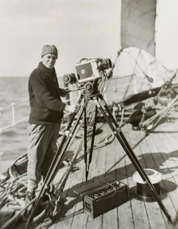 Photo of Frank Hurley on the ship's deck with his camera mounted on a tripod