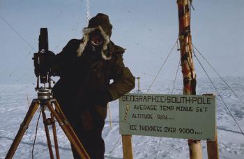 Photo of Director John Shaw at the South Pole with a sign reading 'Geographic South Pole'.