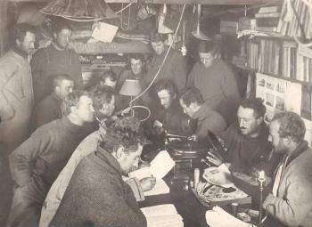 Photo of numerous men cramped in a hut in Antartica around a table reading and writing