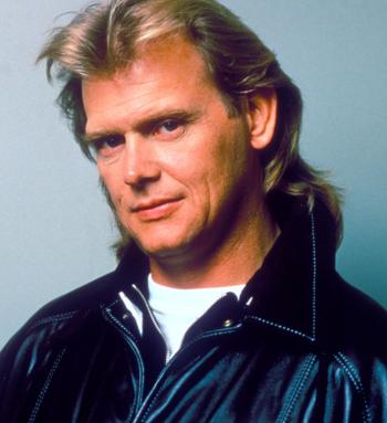 Head and shoulders shot of John Farnham wearing a black leather jacket looking at camera with head slightly tilted.