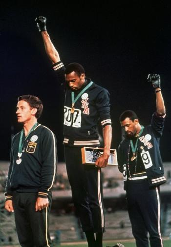 Upper body shot of the three athletes on winners podium. Peter Norman with Tommie Smith and John Carlos on right with gloved fists raised in a salute