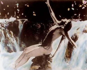Annette Kellerman in a hand-tinted photograph. She appears to be naked but is covered by her long hair. She is sitting on a rock near a waterfall.