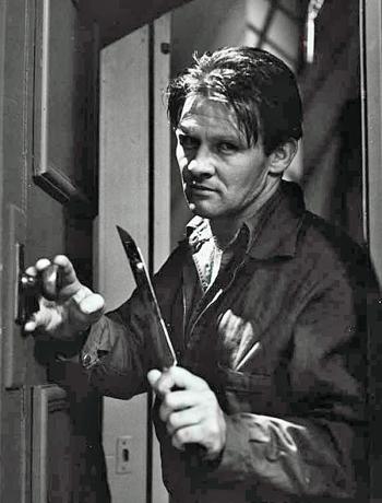 A menacing-looking man pictured from the waist up standing in a doorway holding a large knife.
