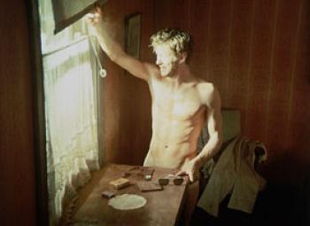 A nude young man stands in a room look out the window into the glaring sunlight