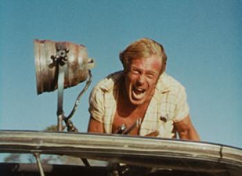 A man screams on top of a car in the arid landscape