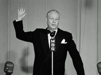Man standing at a microphone in a studio with one hand raised.