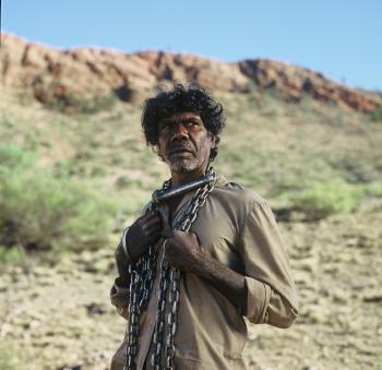 An aboriginal man stands chained in an arid landscape.