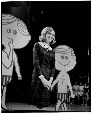 Singer Olivia Newton-John on stage in a TV studio in an episode of the Go Show. She is pictured full length, standing between two lifesize cartoon props of boys wearing short pants.
