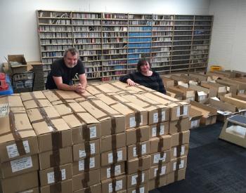 NFSA sound curators resting on a stack of boxes with a shelf of CDs behind them.