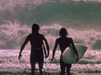 Two surfers holding their surfboards and standing before large waves.