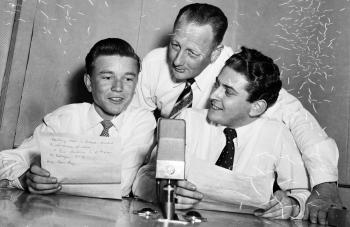 Three men sitting around a radio microphone.