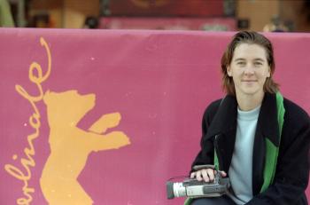 A young woman in front of a pink banner holding a hand-held video camera.