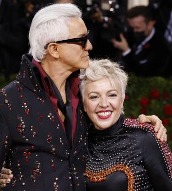 Director Baz Luhrmann wearing sunglasses and an embroidered jacket standing next to Catherine Martin with his arm around her shoulder. She is smiling at the camera. There is a photographer standing in the background.