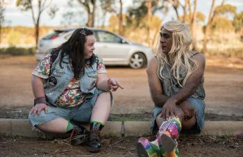 A young woman with Down Syndrome sits beside a drag queen on the ground in front of a parking lot.