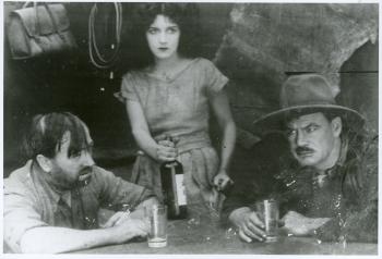 Arthur Tauchert (Jack Bronson) drinking with Charles O'Mara (Ferris), Doris Ashwin (Dell Ferris) is standing beside the table holding a bottle in a film still from The Moth of Moonbi (1926, Charles Chauvel). 