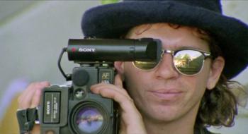 INXS singer Michael Hutchence, wearing reflective sunglasses and a pork pie hat. He is filming with a handicam camera pressed up again his eye.
