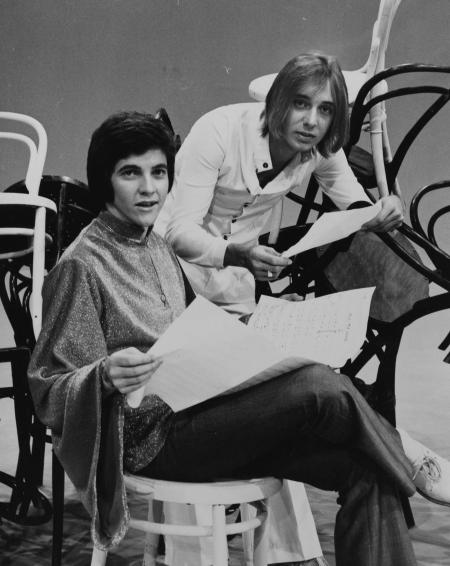 Singer-host Jeff Phillips and singer Russell Morris confer on the set of 1970 pop TV show Sounds Like Us. Phillips is seated and looking over pages of sheet music with Morris leaning on his shoulder. Both are looking at the camera and they are surrounded on the studio floor by a sculptural arrangement of chairs