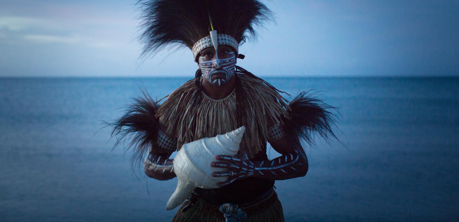 Image featuring Indigenous Francis Williams of the Naygayiw Gigi Dance Troupe in costume standing in front of the sea