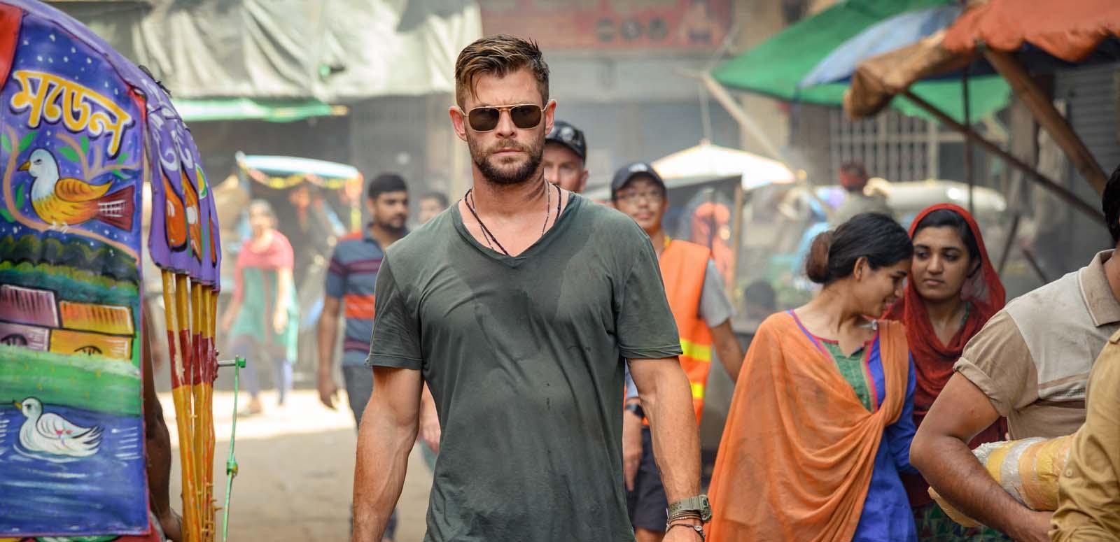 A caucasian assassin in t-shirt and sunglasses walks through a busy market in Bangladesh