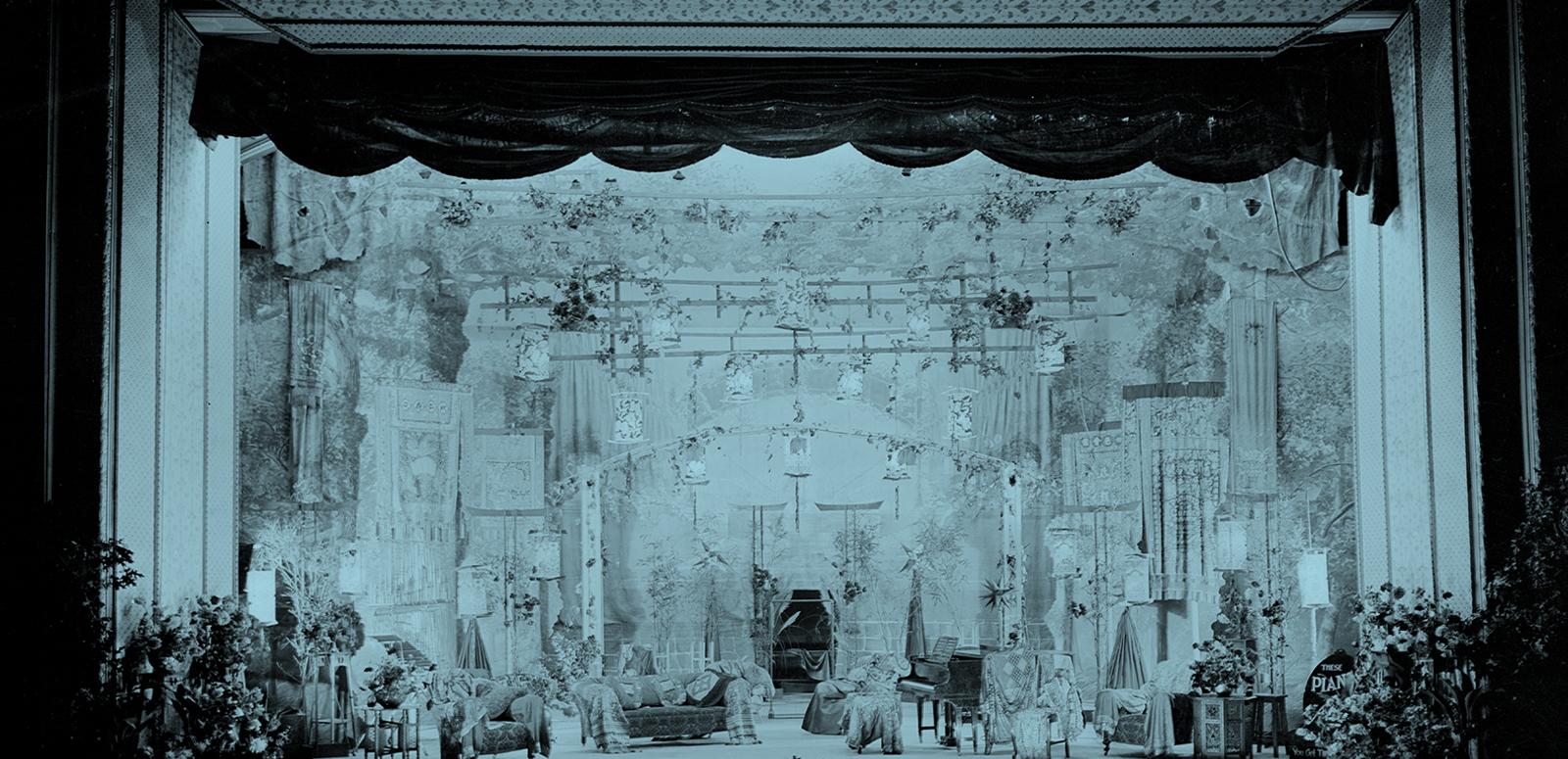 A tinted image of an early 20th century stage before a Corricks family musical performance. It features a piano, an array of chairs and armchairs for the performers, and decorative flowers both on the stage and suspended above it.
