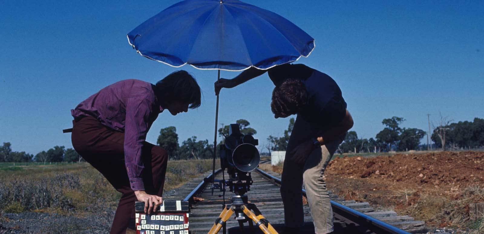 A man holds an umbrella over a film camera positioned on railway tracks while another man holds a clapperboard