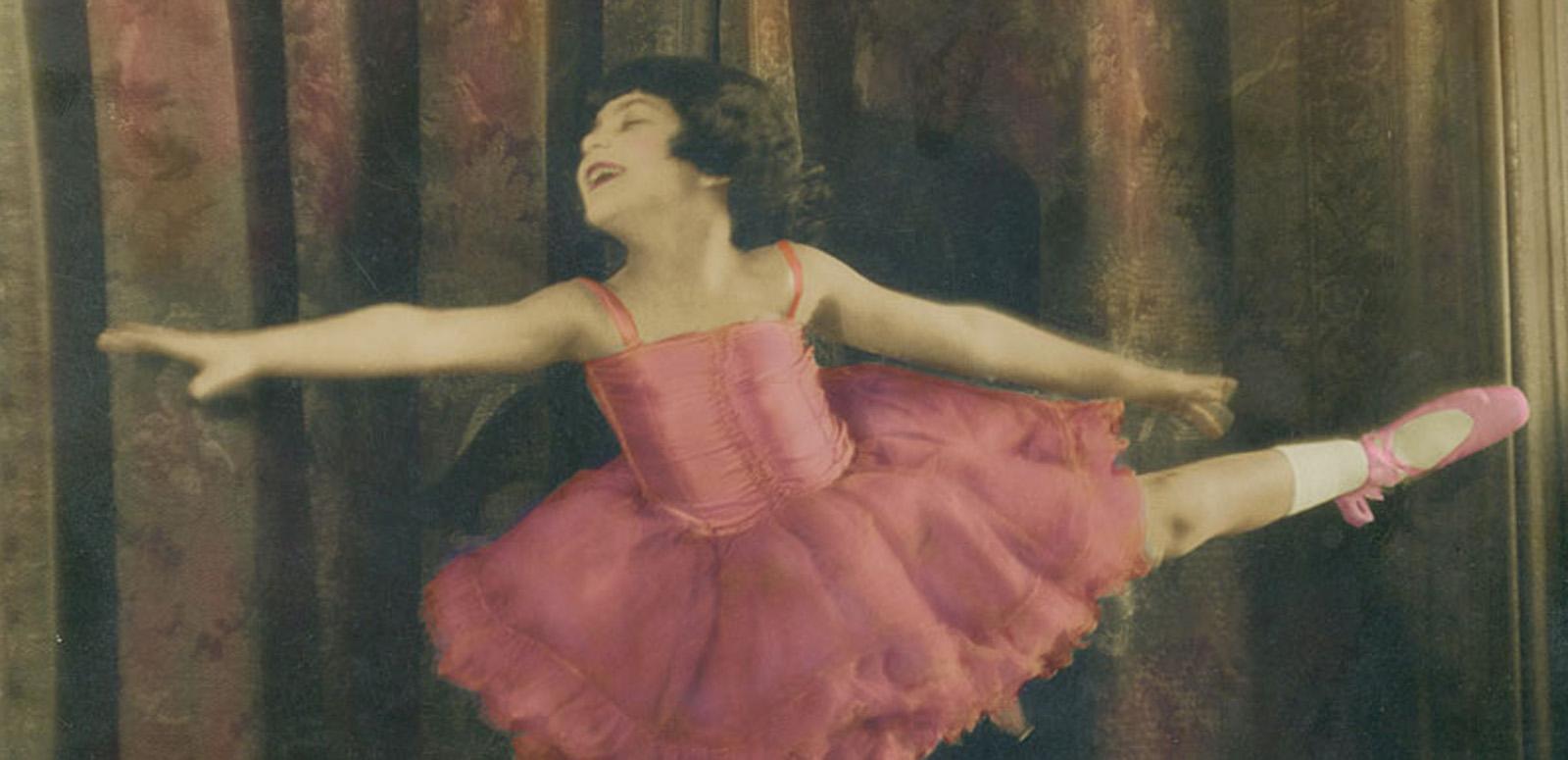 A hand-coloured still of Annette Kellerman as a child. She is dressed in a pink ballet tutu with ballet slippers and is striking a pose with her arms and on leg out.