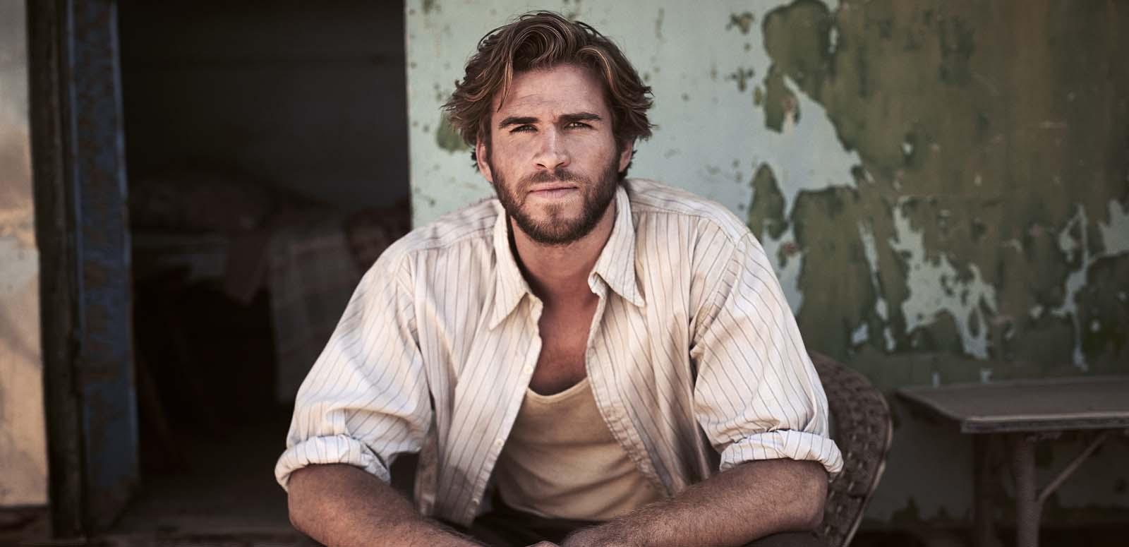 Liam Hemsworth in The Dressmaker, seated in a chair on a verandah in front of a faded wall