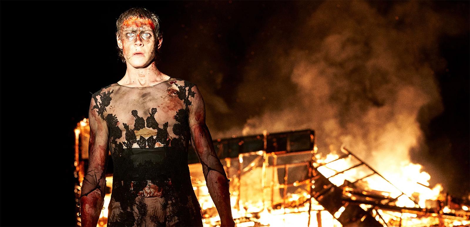 A young man covered in ash and blood standing in front of a house on fire at night
