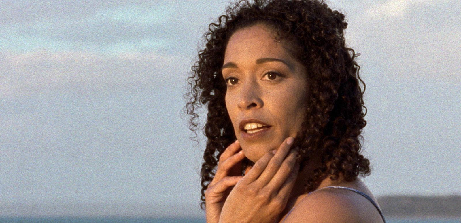 Close up of actor Rachel Maza looking to her left with her hands to her face in a scene from Radiance. The ocean and a cloudy sky can be seen in the background.