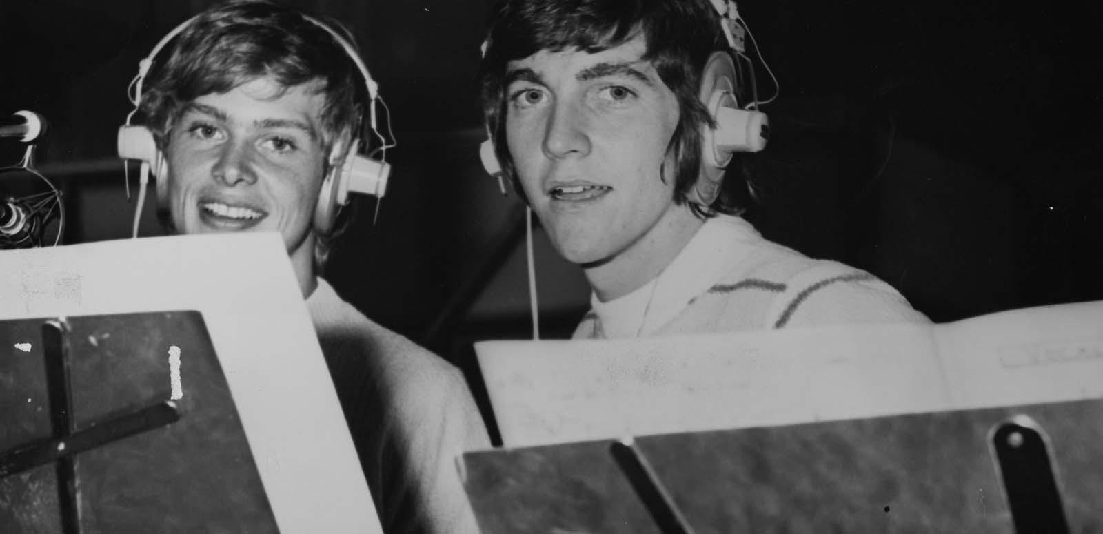 John Farnham and Jeff Phillips wearing headphones and standing behind music stands during a session in the recording studio in 1970