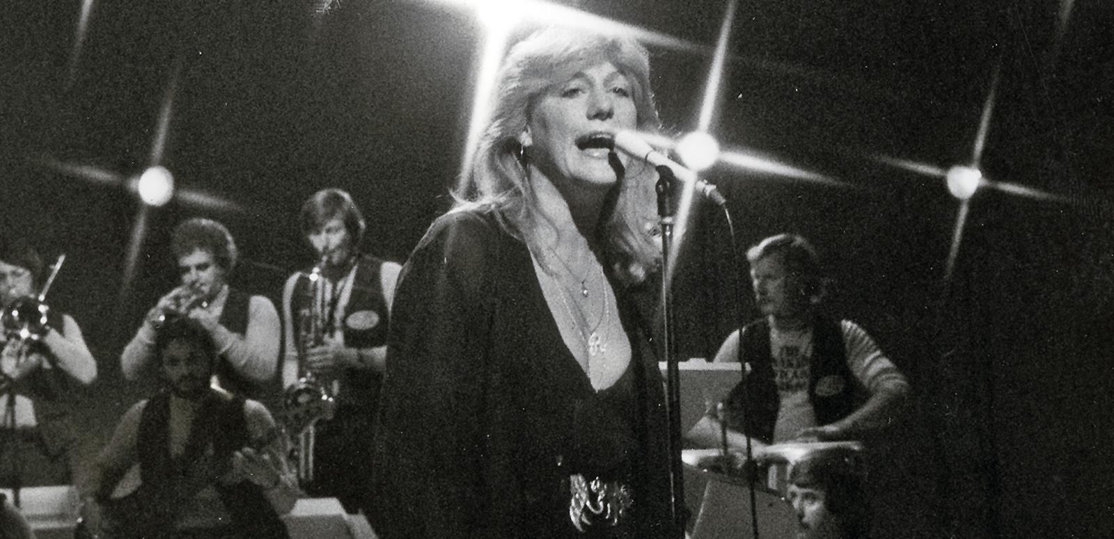 Singer Renee Geyer pictured from the waist up, singing into a microphone with band members in the background behind her.