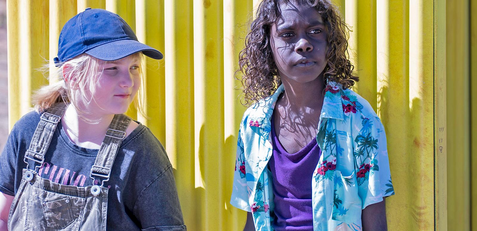 Two kids standing side by side against a yellow corrugated iron fence. One is white girl with straight blond hair in a ponytail, wearing denim overalls and a cap, and the other is a First Nations boy with curly dark hair wearing a purple singlet and blue Hawaiian shirt.